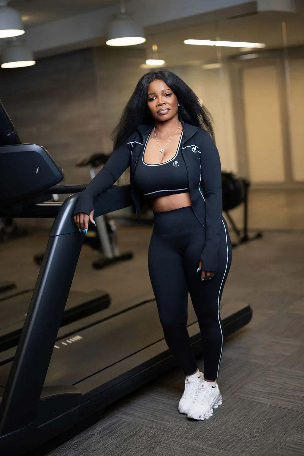 Woman in black athleisure set with white sneakers standing by treadmill in gym