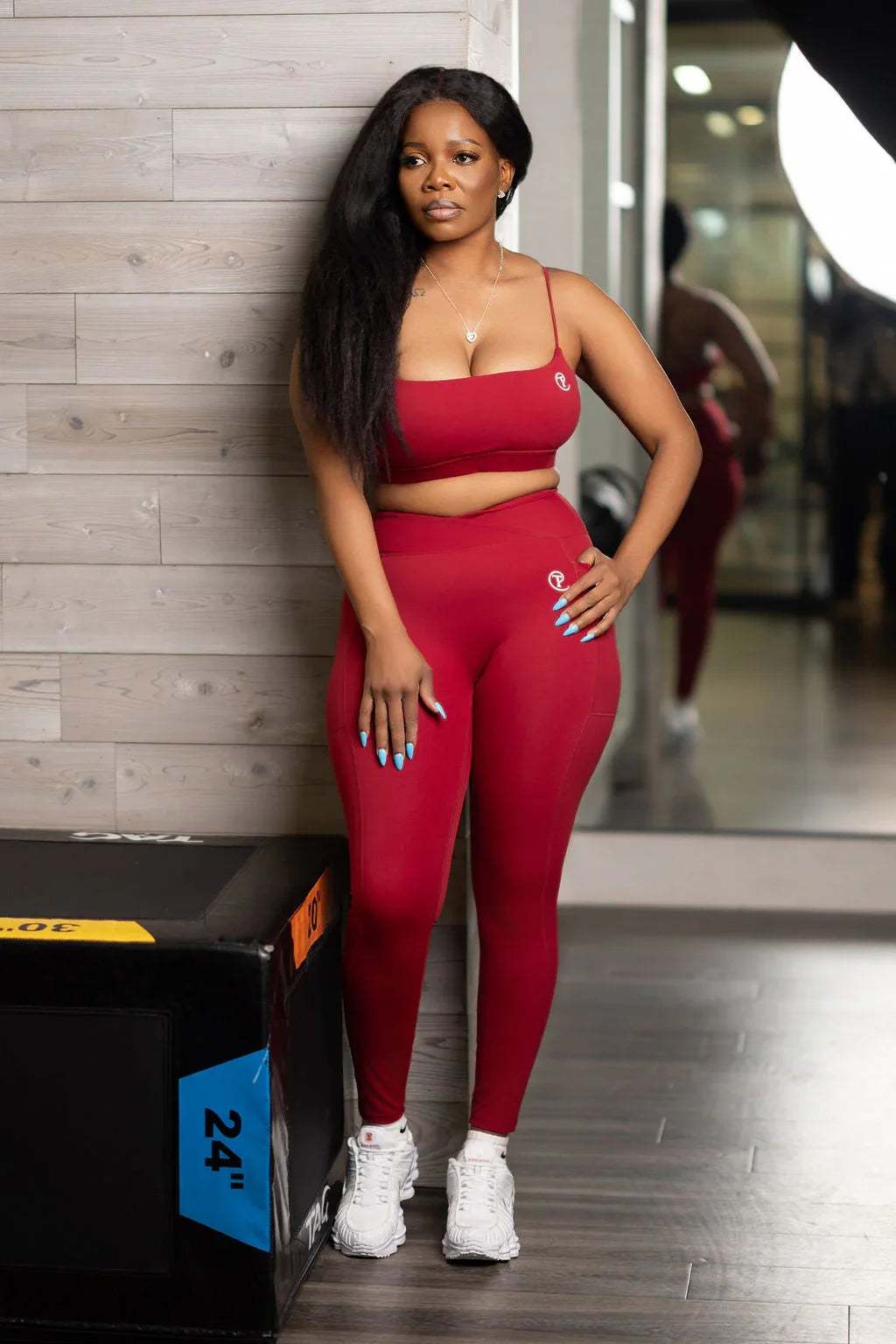 Woman in burgundy sports bra and leggings set, posing in gym with exercise box, wearing white sneakers.