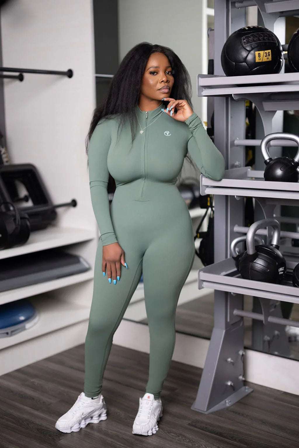 Woman in sage green full-body athletic jumpsuit posing confidently in a modern gym