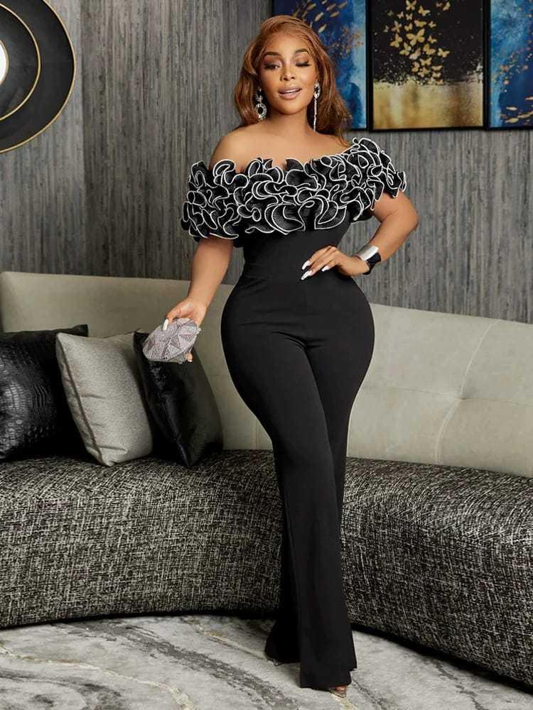 Woman in stylish black off-shoulder jumpsuit with ruffled neckline, holding clutch, in modern living room