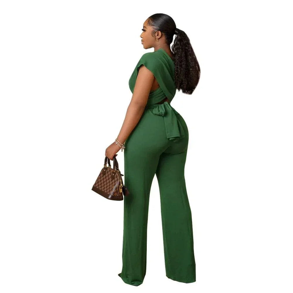 Woman in a stylish green jumpsuit with a tie-back, holding a brown patterned handbag.