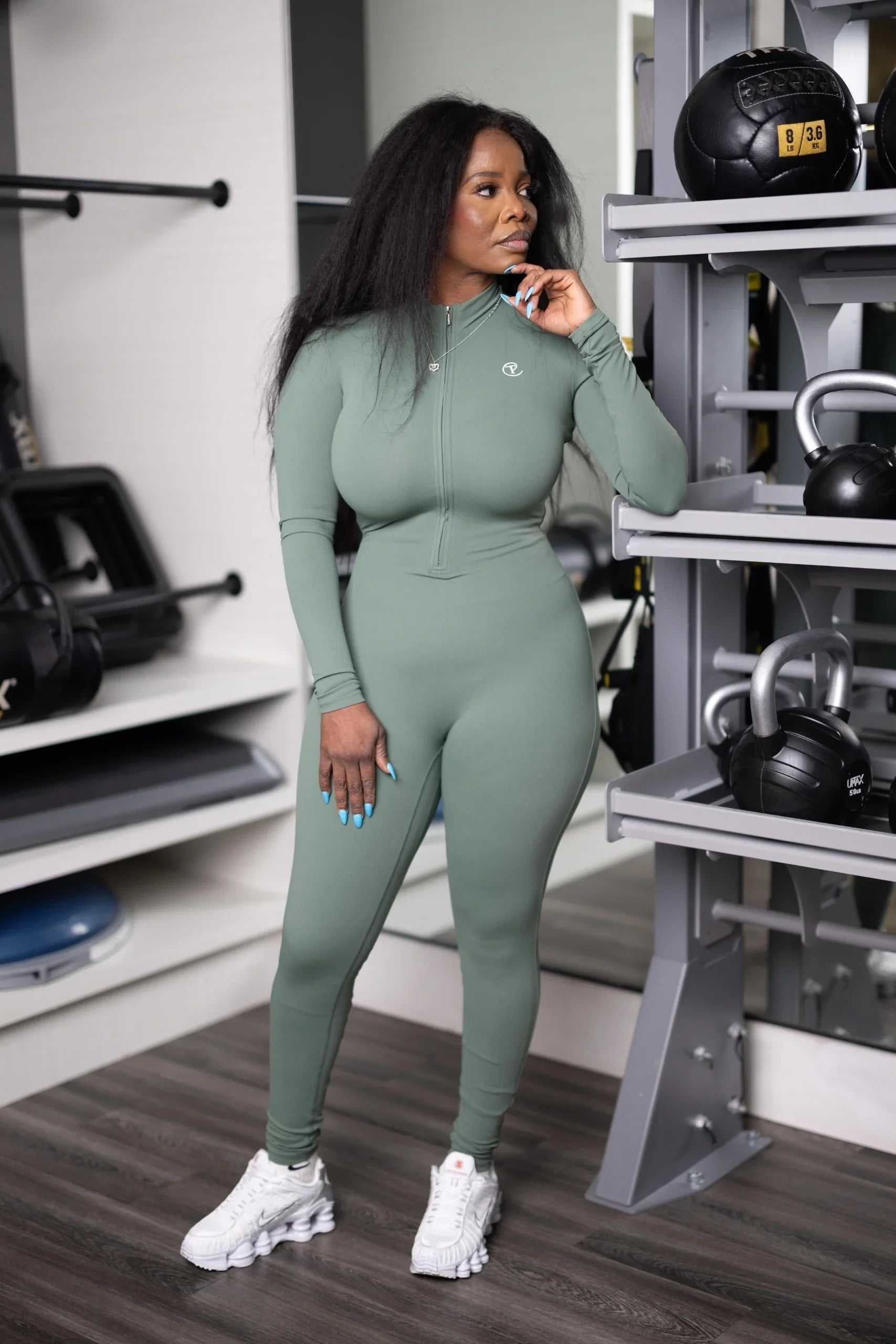 Woman in sage green zip-up athletic jumpsuit and white sneakers standing in gym with weights