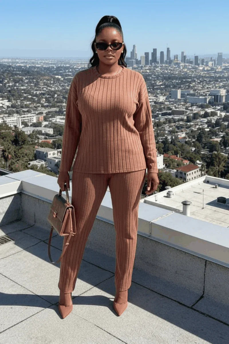 Woman in ribbed brown sweater and pants, heels, holding handbag, cityscape background