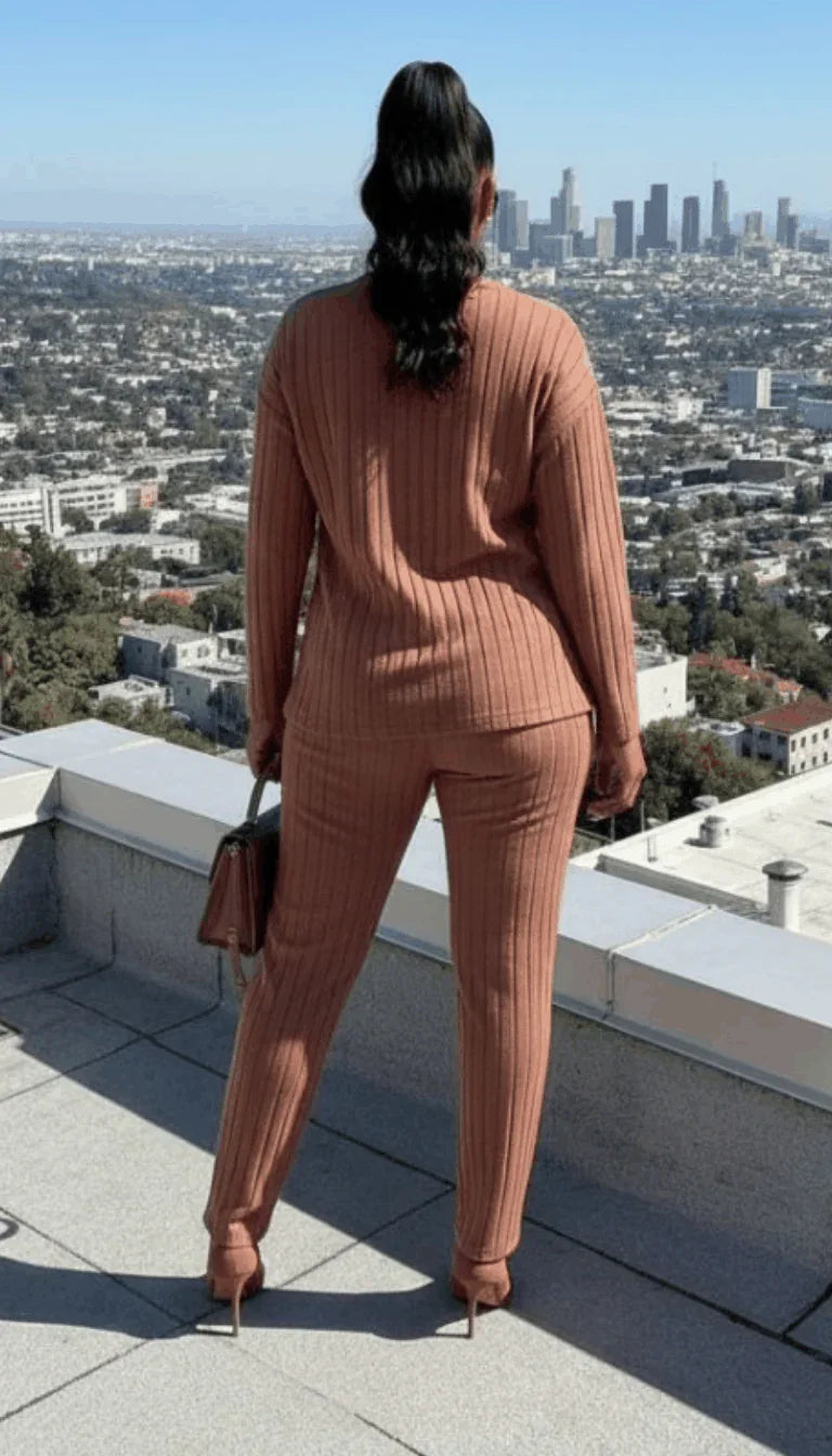 Woman in ribbed, brown knit pantsuit and heels overlooking city skyline from rooftop